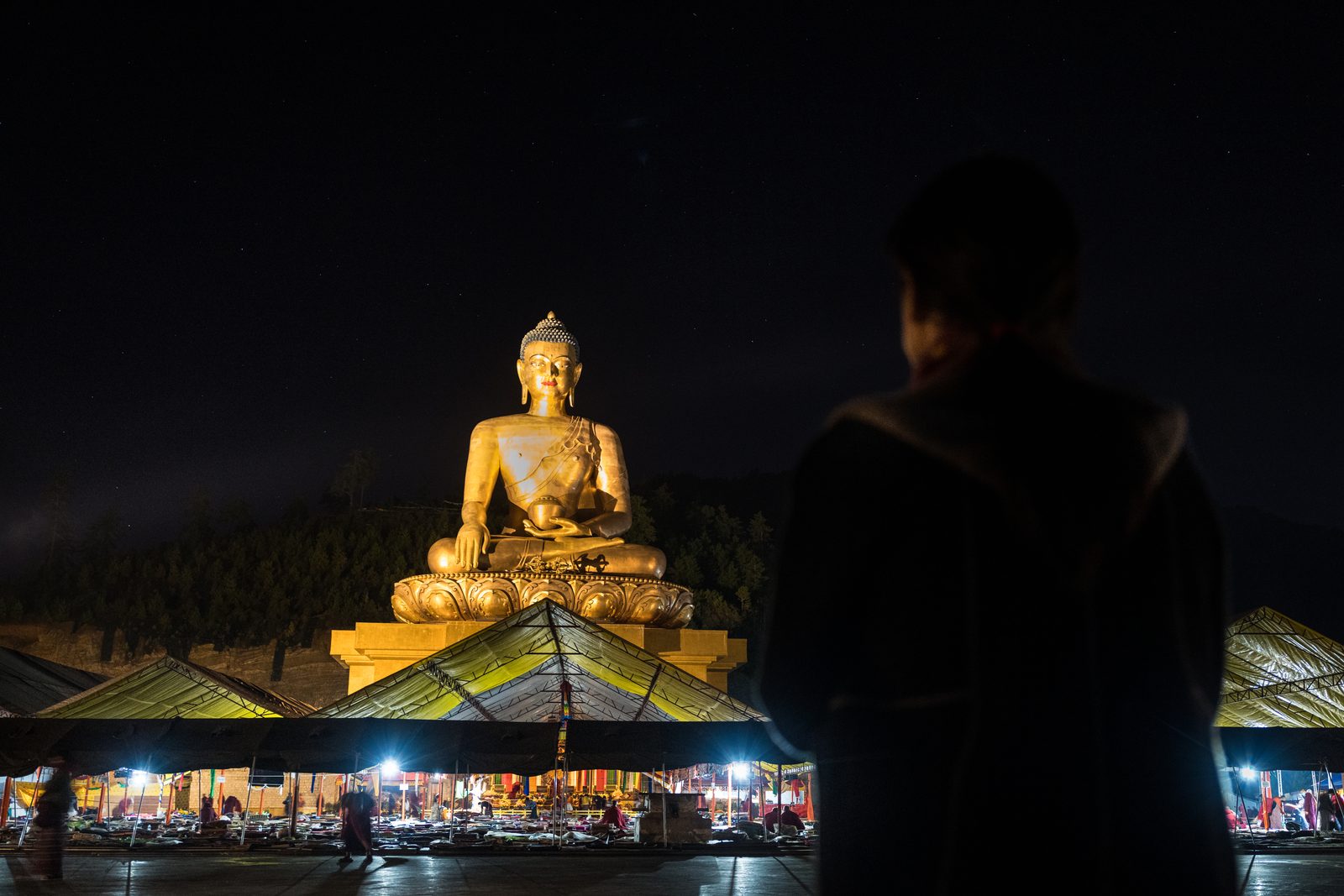 The gilded Buddha Dordenma lit at night above Thimphu, with festival tents below and a visitor watching in silhouette