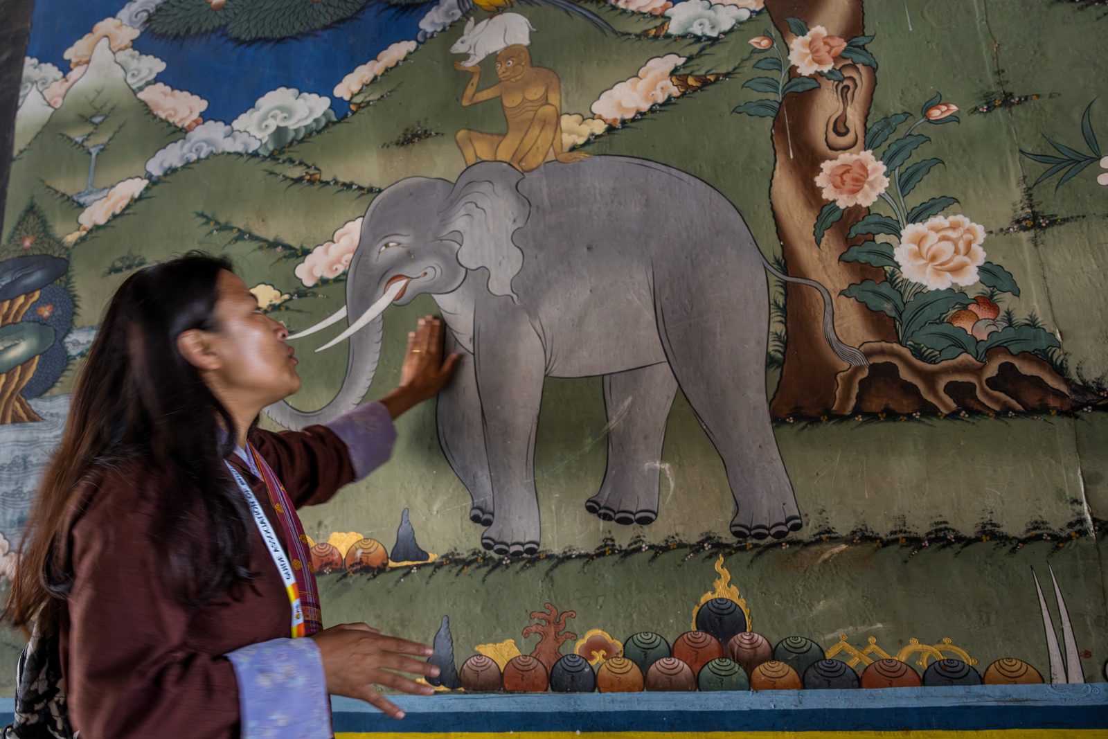 A Bhutanese guide with her ID lanyard points out a temple mural of the Four Harmonious Friends, an elephant carrying a monkey and a hare