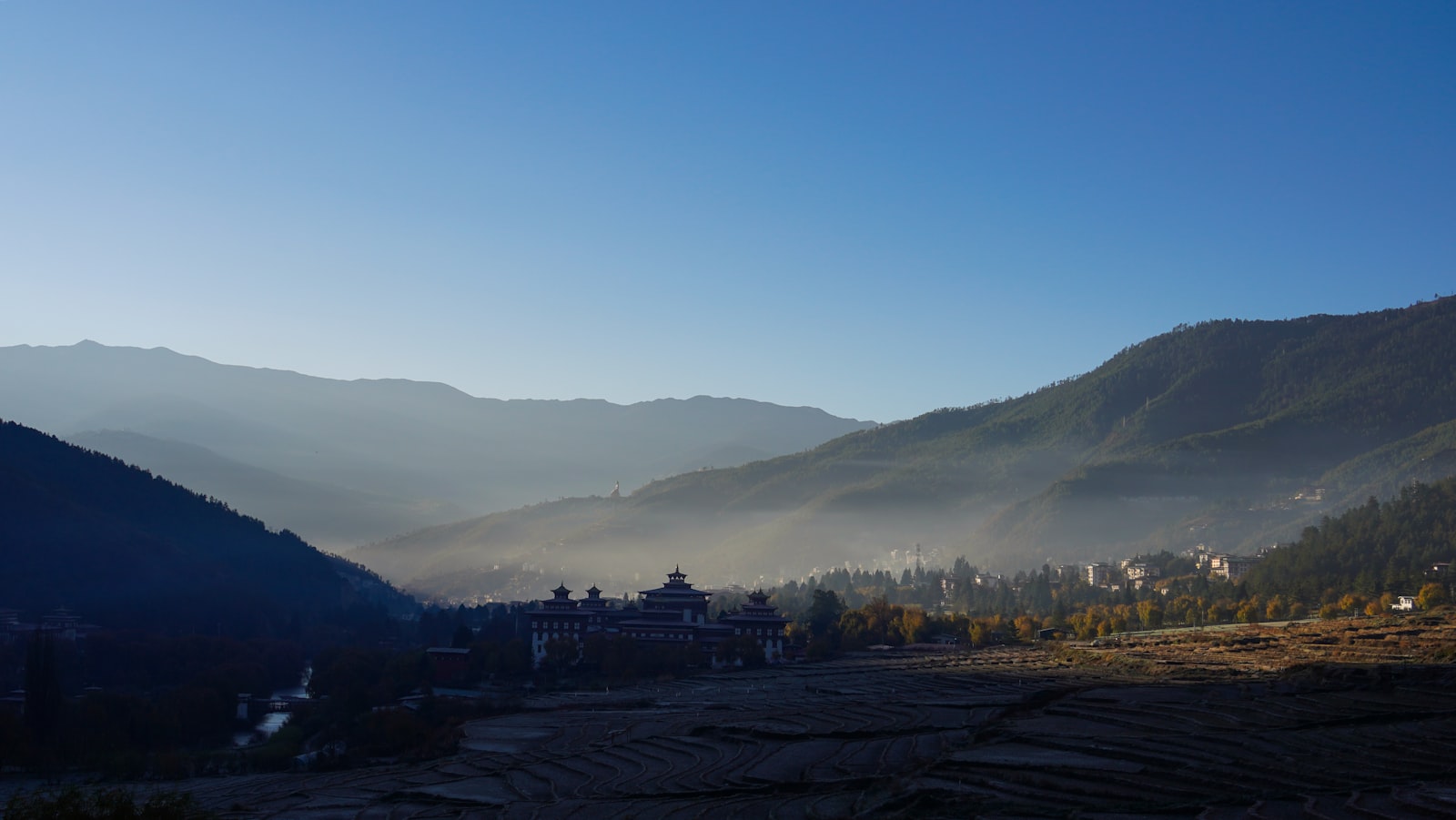 A misty Bhutanese valley at sunrise with a monastery below the ridge