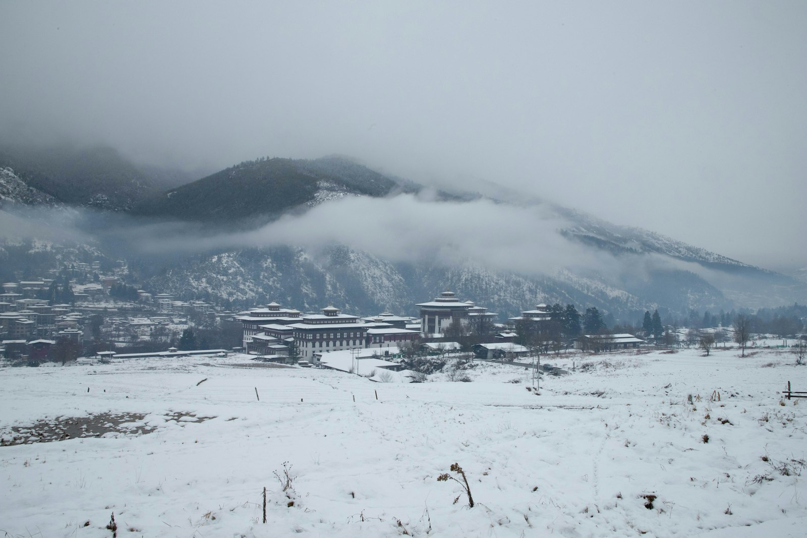 Tashichho Dzong across snow-covered fields in winter, cloud low on the mountain